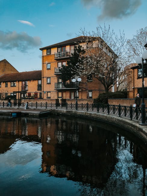 A modern multi-story residential building situated alongside a river, with a facade composed of dark brickwork and tall, narrow windows. The property features a stepped design with varying levels, pro